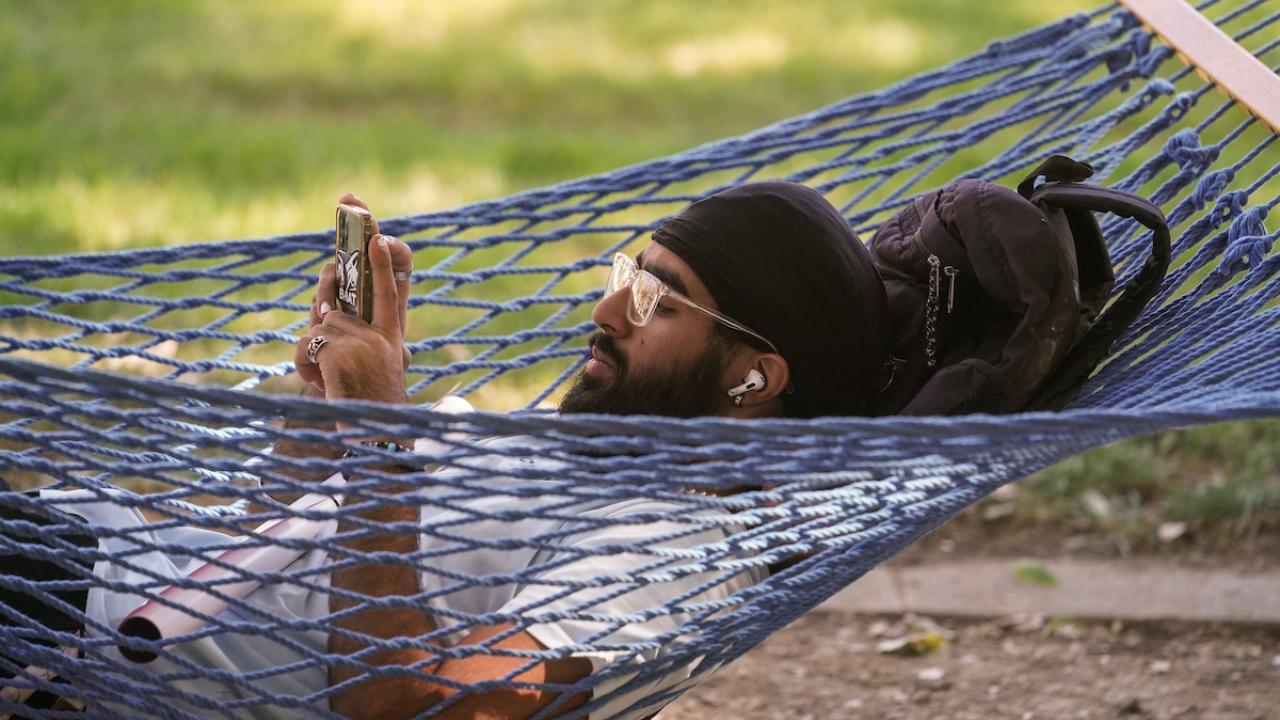 A student uses his cell phone while resting in a hammock on the quad.