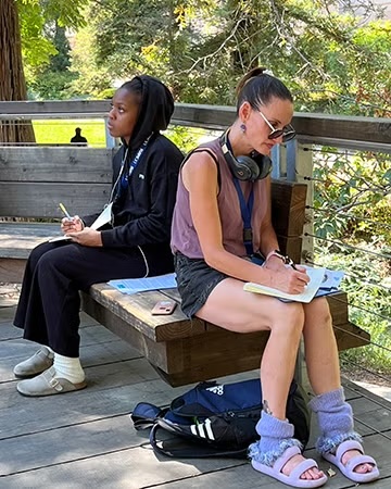 A student sits in a shaded outdoor area of the arboretum, writing in a notebook while surrounded by greenery. A backpack rests nearby, and the atmosphere is calm and focused.