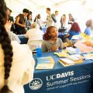 A lively informational fair under a large tent, where students engage with representatives at various tables. In the foreground, a woman behind a UC Davis Summer Sessions table speaks with a student holding informational flyers. The table is covered with brochures, flyers, and giveaways. Other tables and attendees are visible in the background, contributing to a busy and energetic atmosphere. The UC Davis Summer Sessions banner includes the logo and reads 'Office of Undergraduate Education.